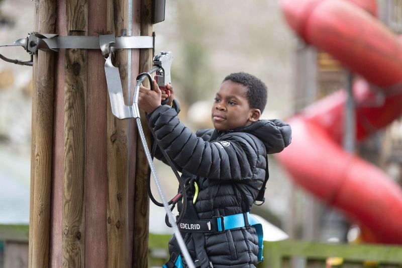 A child dressed in a black padded jacket and safety harness is attaching a carabiner to a metal fixture on a wooden pole at an outdoor adventure park. A red spiral slide is visible in the background.
