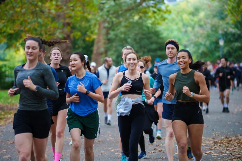 A group of runners in athletic wear take part in a communal run through a park, surrounded by trees and greenery.