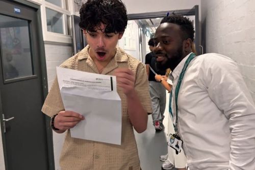 Two individuals stand in a hallway with white walls and doors on either side. One person holds a piece of paper, while the other wears a lanyard with an ID badge and stands beside him.