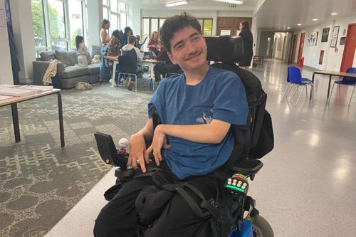 A person in a blue t-shirt sits in a wheelchair smiling at the camera inside a spacious room with large windows. In the background, groups of people are gathered around tables and sofas, engaged in conversation or working.