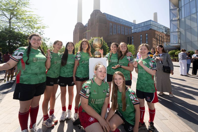 A group of young female rugby players standing around the new Women's Rugby World Cup trophy in front of Battersea Power Station