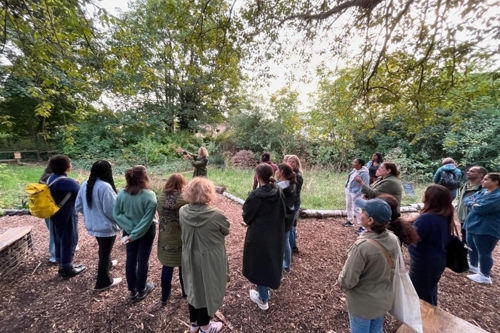 A group of women listen to a speaker in a garden surrounded by trees